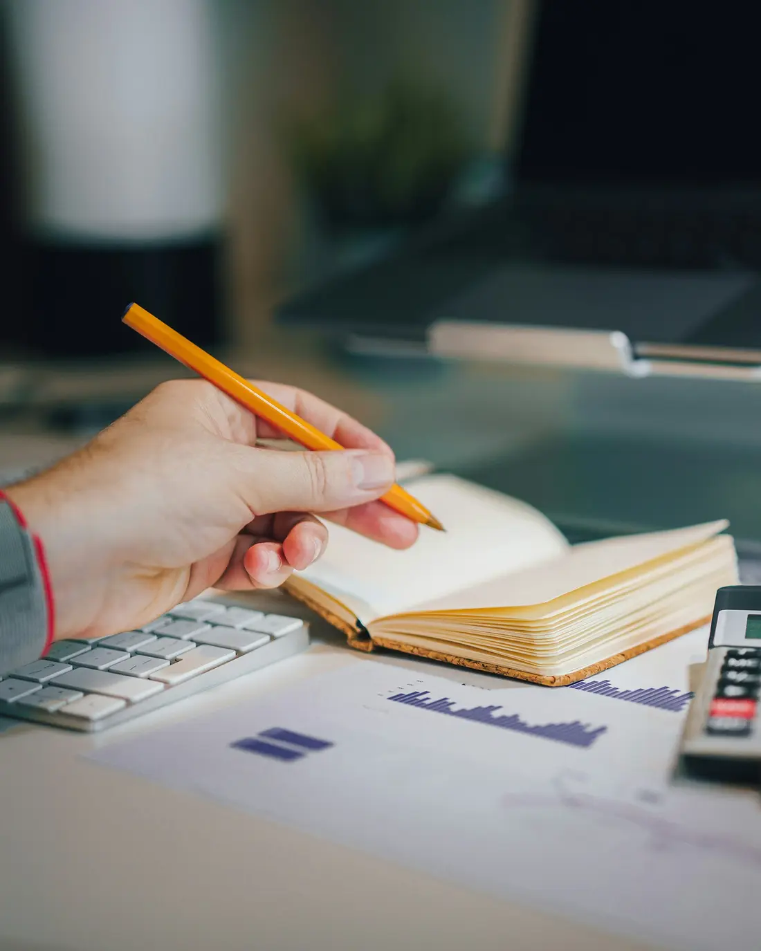 Mão com lápis sobre um caderno, ao lado de calculadora e gráficos impressos
